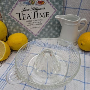 May include: A clear glass lemon squeezer with a ridged rim and a central cone. The squeezer is on a blue and white checked tablecloth. There are lemons and a white jug in the background.