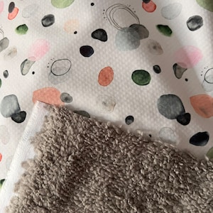 Könnte beinhalten: Ein weißer Stoff mit einem Muster aus abstrakten Formen in Rosa-, Grün-, Orange- und Grautönen. Der Stoff ist mit einem weichen, grauen, flauschigen Material kombiniert.