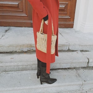 May include: A woman wearing a red coat and black boots is carrying a woven straw bag with a long strap.