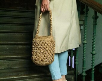 May include: A woman wearing a beige trench coat and blue jeans is holding a brown crocheted bag. She is standing on a set of wooden stairs with a green metal railing.