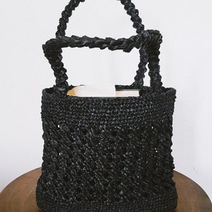 May include: A black woven tote bag with a thick, twisted rope handle. The bag has a diamond-shaped pattern and is made of a natural material.