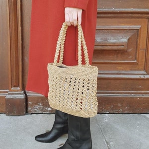 May include: A tan crocheted tote bag with a woven design. The bag has two handles and is being held by a person wearing a red coat and black boots.