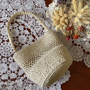 May include: A cream-colored crocheted tote bag with a woven handle. The bag is sitting on a white crocheted doily.