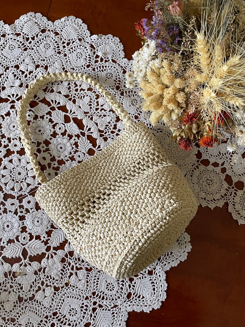 May include: A cream-colored crocheted tote bag with a woven handle. The bag is sitting on a white crocheted doily.