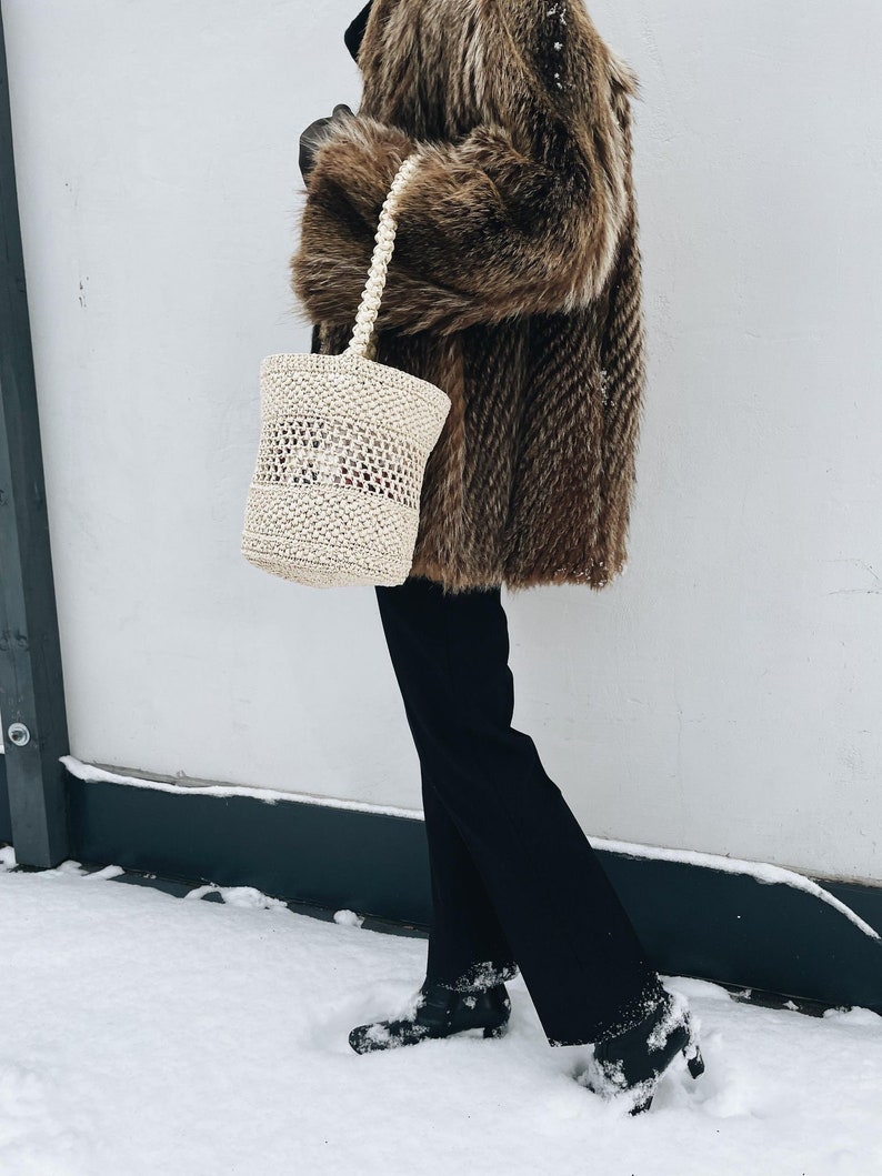 A female silhouette dressed in vintage fur coat, holds a straw-coloured basket in her left arm. Wearing black trousers and black shoes covered thinly in snow. She stands against a white wall, there is a thin covering of snow on the ground.
