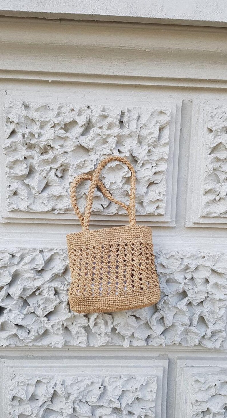 May include: A small, beige, crocheted tote bag with a woven design hangs against a white textured wall.