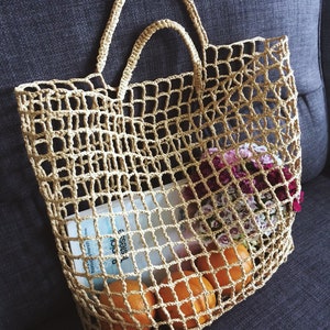 May include: A beige, open-weave tote bag filled with oranges, a book, and flowers. The bag has two handles and is sitting on a dark gray surface. The bag's design allows the contents to be visible.