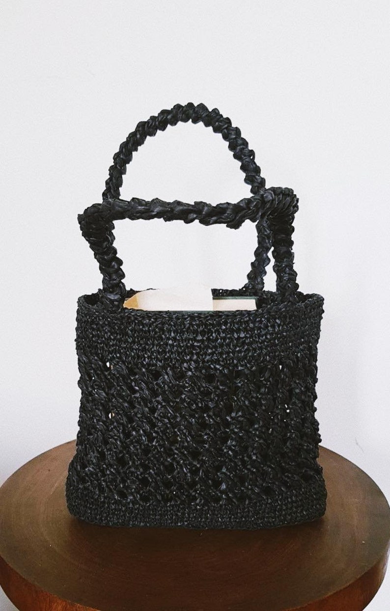 May include: A black woven tote bag with a thick, twisted rope handle. The bag has a diamond-shaped pattern and is made of a natural material.