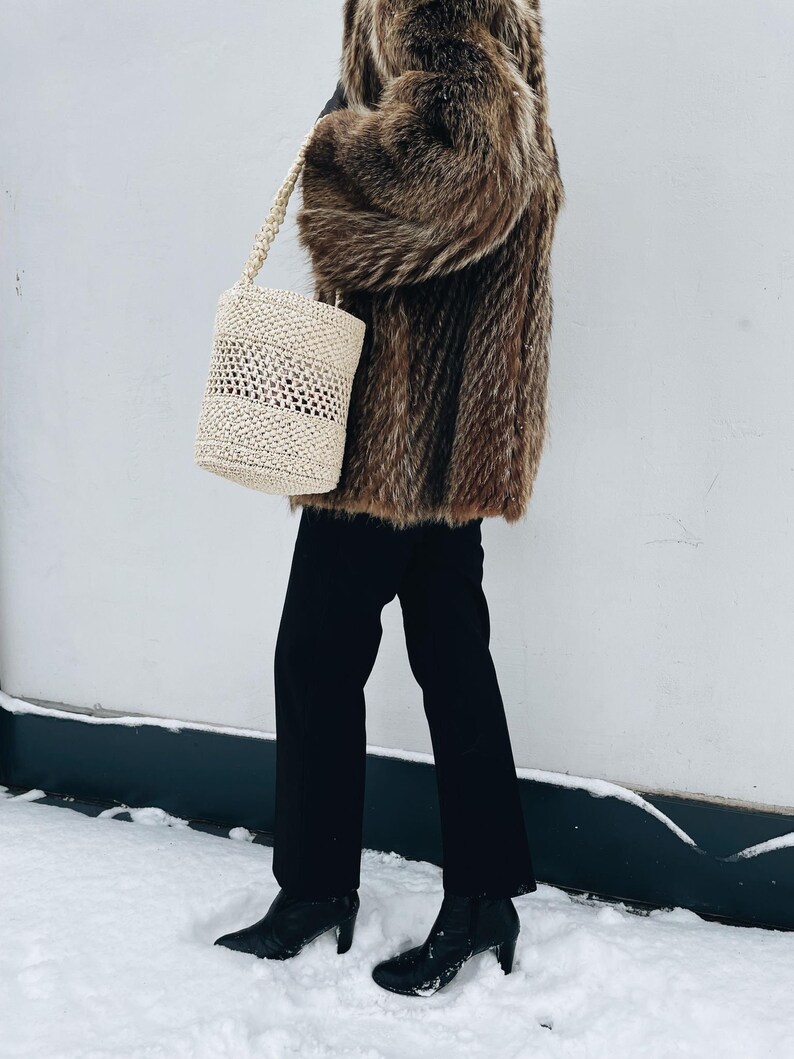 May include: A person wearing a brown fur coat, black pants, and black heeled boots is holding a white woven tote bag. The person is standing in the snow.