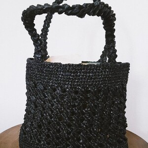May include: A black woven straw tote bag with a thick rope handle. The bag has a diamond pattern and is made of a lightweight material.