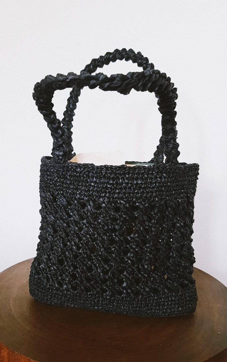 May include: A black woven straw tote bag with a thick rope handle. The bag has a diamond pattern and is made of a lightweight material.