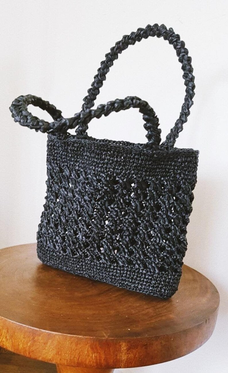 May include: A black woven tote bag with a braided handle. The bag has a square shape and a delicate open weave pattern.
