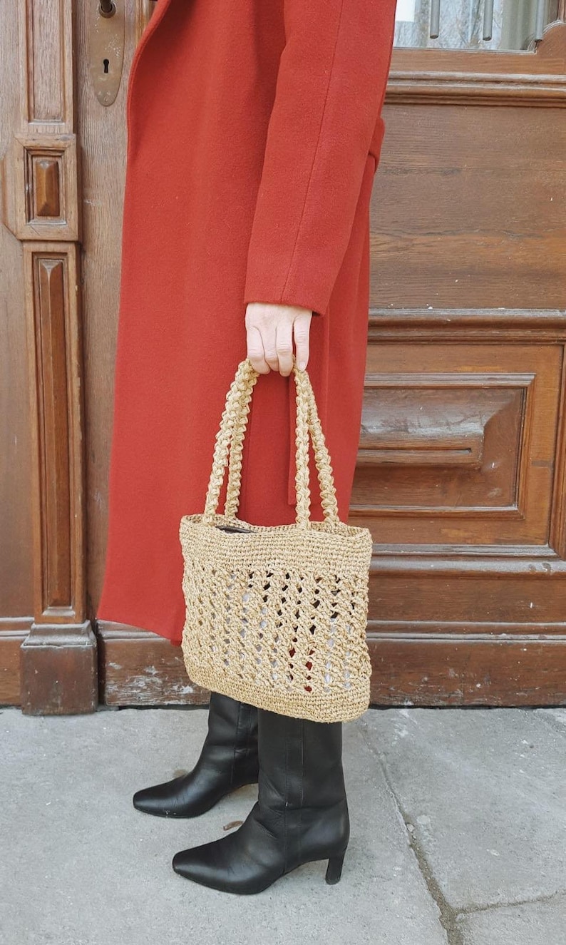 May include: A tan crocheted tote bag with a woven design. The bag has two handles and is being held by a person wearing a red coat and black boots.