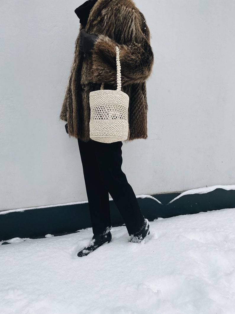 May include: A white crocheted tote bag with a thick rope handle. The bag is being held by a person wearing a brown fur coat and black pants.