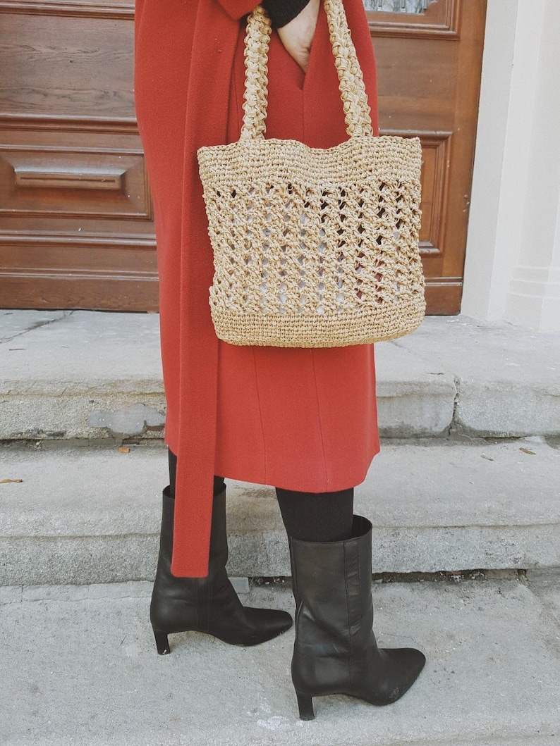 May include: A woman wearing a red coat and black boots is carrying a woven straw tote bag with a wide, open top.