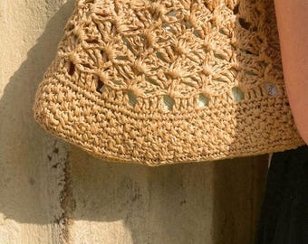 May include: A woven raffia tote bag with a floral pattern. The bag is a light tan color and has a braided handle. The bag is held against a textured beige wall.