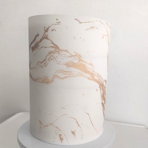 Può includere: Una torta bianca con un design dorato effetto marmo, esposta su un'alzatina bianca con base dorata. La torta cilindrica presenta un motivo dorato fluido e organico.