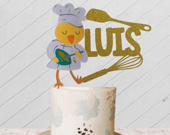 May include: A white cake with a silver and pink design topped with a yellow chick wearing a chef's hat and holding a mixing bowl. The chick is holding a spoon and there is a gold glitter spatula and whisk behind it. The cake topper also includes the name "LUIS" in gold glitter.