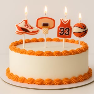 May include: A white iced cake decorated with orange frosting and basketball-themed candles. The candles include a basketball shoe, a hoop, a jersey with the number 33, and a basketball. The candles are lit with flames.
