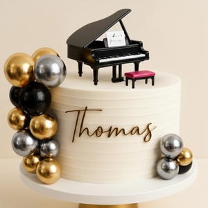 Può includere: Una torta bianca decorata con un pianoforte a coda nero, una panchina rosa e decorazioni con palloncini dorati, argentati e neri. Il nome "Thomas" è scritto sul lato della torta. La torta è su un vassoio bianco.