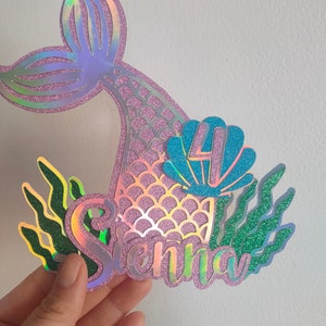 May include: A mermaid-themed cake topper with a holographic tail, the name "Sienna", and the number "4" inside a blue seashell. Green glittery seaweed accents the design. The topper is held by a hand.