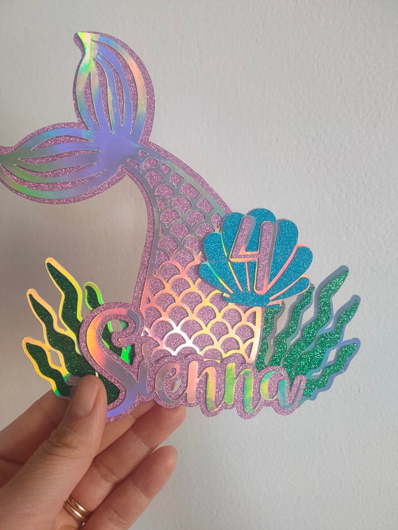 May include: A mermaid-themed cake topper with a holographic tail, the name "Sienna", and the number "4" inside a blue seashell. Green glittery seaweed accents the design. The topper is held by a hand.