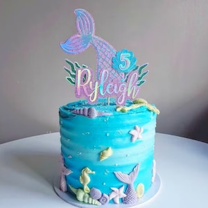 May include: A round, blue frosted cake with a mermaid theme. The cake is decorated with edible seashells, starfish, and seahorses. A holographic mermaid tail cake topper with the name "Ryleigh" and the number "5" is on top.