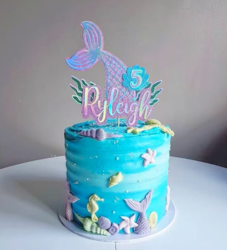 May include: A round, blue frosted cake with a mermaid theme. The cake is decorated with edible seashells, starfish, and seahorses. A holographic mermaid tail cake topper with the name "Ryleigh" and the number "5" is on top.