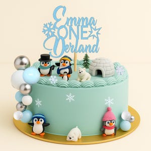 May include: A light blue frosted cake with winter-themed decorations. The cake features penguin figurines, a polar bear, an igloo, and a cake topper that reads "Emma ONE Verland". Silver and blue balloon accents surround the cake.
