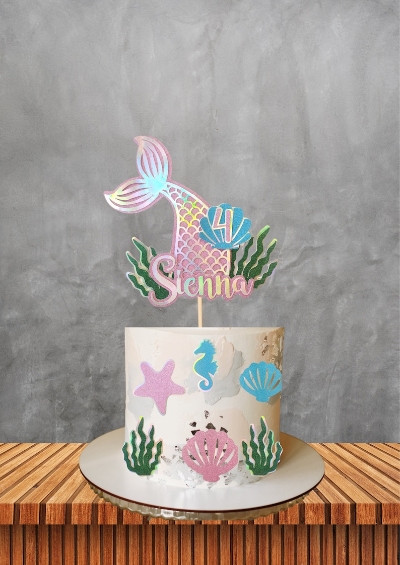 May include: A white frosted cake decorated with mermaid-themed cutouts, including a starfish, seahorse, and seashells. A mermaid tail cake topper with the name "Sienna" and the number "4" is placed on top. The cake sits on a wooden table.