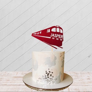 Op de afbeelding: Een witte cake met een rode trein topper waarop "JASPER" staat. De cake is versierd met een witte en grijze frosting.