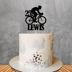 May include: Black silhouette cake topper of a cyclist riding a bicycle with the number 25 and the name Lewis. The topper is on a white cake with a light pink and gray frosting.