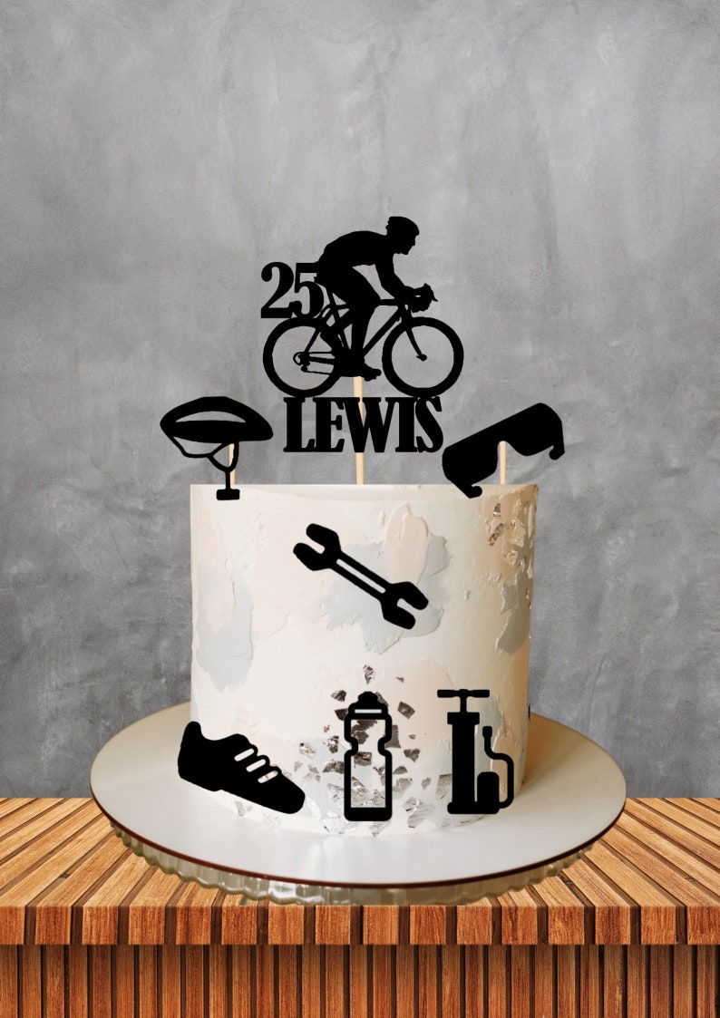 May include: Black and white silhouette cake topper featuring a cyclist riding a bicycle with the number 25 and the name Lewis. The cake topper is surrounded by other cycling-themed decorations, including a helmet, sunglasses, a wrench, a water bottle, and a bicycle pump.