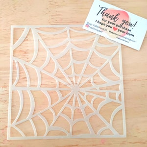 Peut inclure: Pochoir en plastique blanc représentant une toile d'araignée. Le pochoir est posé sur une surface en bois.