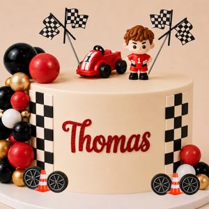 Può includere: Una torta rotonda con glassa beige liscia, decorata con un'auto da corsa rossa, una piccola figura in tuta da corsa rossa e bandiere a scacchi. Il nome Thomas è scritto in rosso. Sfere nere, rosse, bianche e dorate decorano i lati della torta.