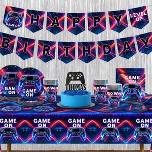 May include: A gaming-themed birthday party set-up with a blue cake, plates, cups, and a banner that reads "Happy Birthday." The decorations feature a neon controller design and the words "Game On" and "Level Up."