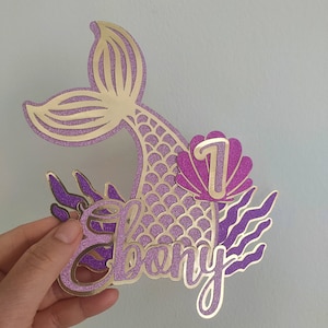 May include: A glittery gold and purple mermaid-themed cake topper. The design includes a mermaid tail, the name "Ebony", the number "1", and decorative seaweed elements. The topper is held by a hand.