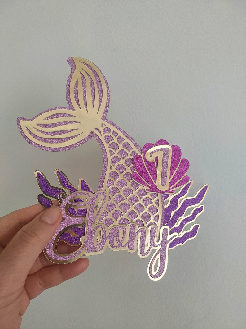 May include: A glittery gold and purple mermaid-themed cake topper. The design includes a mermaid tail, the name "Ebony", the number "1", and decorative seaweed elements. The topper is held by a hand.