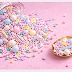 Peut inclure: Un pot renversé de vermicelles colorés et une cuillère dorée remplie de vermicelles sur une surface rose. Les vermicelles comprennent des sphères pastel, des formes de fleurs et des bonbons en forme de bâtonnets, créant un affichage festif.