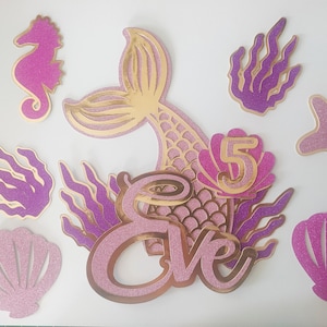 May include: A collection of mermaid-themed party decorations. Includes a mermaid tail, seahorse, starfish, seaweed, and seashell cutouts in pink and gold glitter. The name "Eve" and the number "5" are also featured.