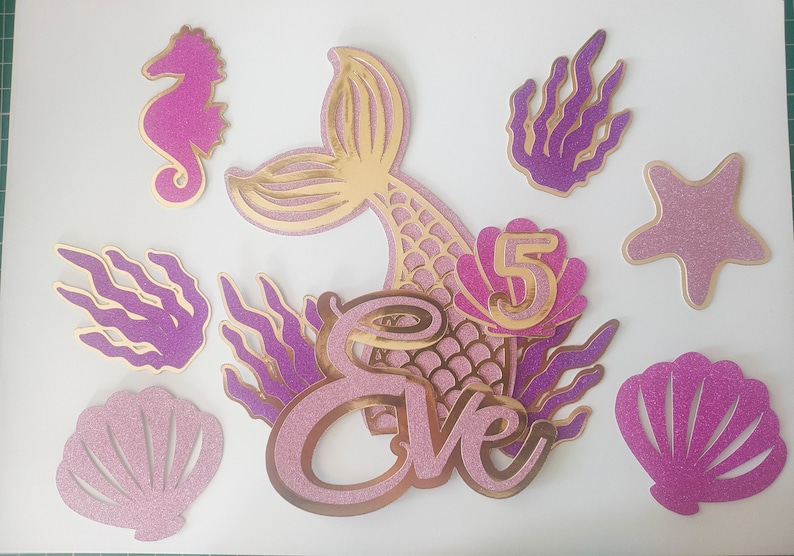 May include: A collection of mermaid-themed party decorations. Includes a mermaid tail, seahorse, starfish, seaweed, and seashell cutouts in pink and gold glitter. The name "Eve" and the number "5" are also featured.