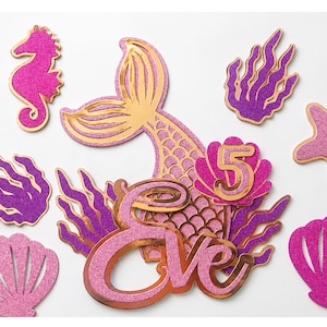 May include: A collection of glittery mermaid-themed party decorations. Includes a mermaid tail, seahorse, starfish, seashells, and seaweed in shades of pink, purple, and gold. The name "Eve" and the number "5" are also featured.
