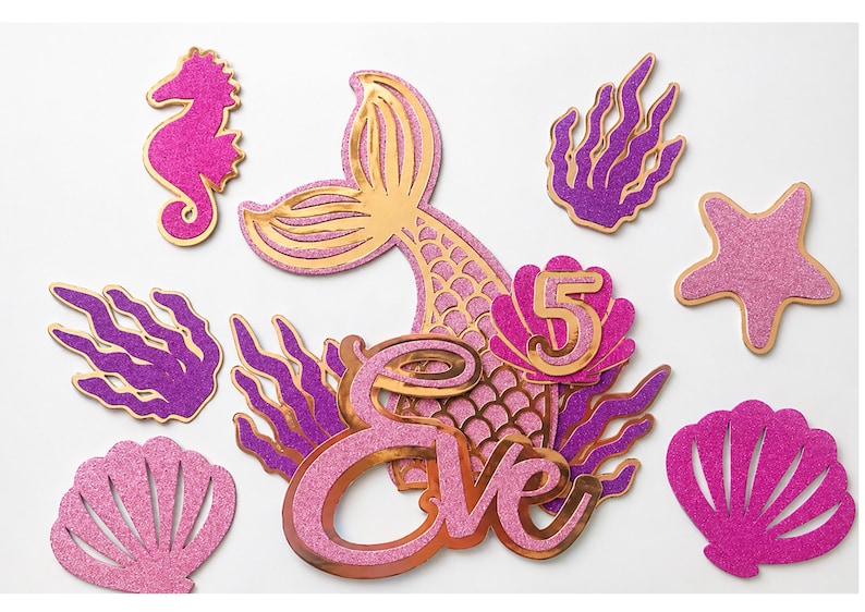 May include: A collection of glittery mermaid-themed party decorations. Includes a mermaid tail, seahorse, starfish, seashells, and seaweed in shades of pink, purple, and gold. The name "Eve" and the number "5" are also featured.