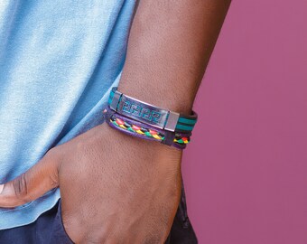 May include: A person wearing a blue t-shirt and black pants with a silver and black bracelet with a pattern and a multi-colored braided bracelet.