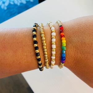 May include: Four beaded bracelets stacked on a wrist. The bracelets are made of small, round beads in black, gold, white, and rainbow colors.