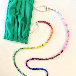 May include: A rainbow beaded mask chain with a green fabric face mask. The chain is made of small, colorful beads and has a white cord with gold clasps.