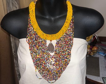 May include: A multi-layered beaded necklace with a wooden pendant. The necklace is made of small, colourful beads and has a yellow beaded collar. The necklace is paired with matching yellow and blue beaded earrings.