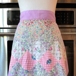 May include: A vintage-style half apron with a lilac waistband and scalloped hem. The apron features a patchwork design with floral and geometric patterns in pink, white, and blue. The apron is displayed on a mannequin.