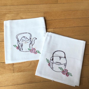 May include: Two white tea towels with embroidered images of teapots and flowers. The teapots are outlined in black and the flowers are red and green.
