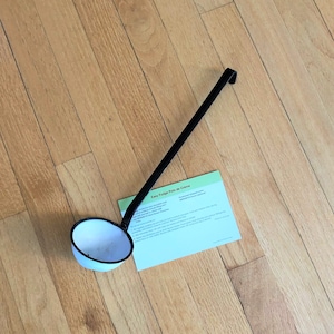 May include: A white enamel ladle with a black handle on a wooden floor. A green and white recipe card for "Easy Fudge Pots de Creme" is lying on the floor next to the ladle.